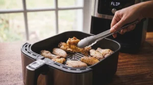 A person uses metal tongs to flip chicken wings inside the basket of a stainless steel air fryer on a wooden countertop, with the digital display reading "FLIP".