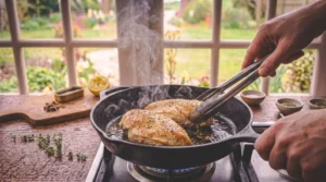 A person uses metal tongs to flip seasoned chicken breasts searing in a hot cast-iron skillet on a stove, creating visible steam. A rustic wooden table with fresh herbs, cut lemon, and small spice bowls sits in front of a bright window.